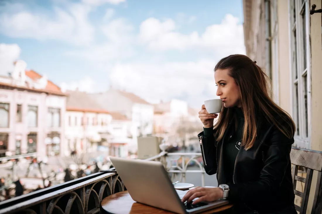 Młoda kobieta pracuje na laptopie i pije kawę na tarasie z widokiem na miasto
