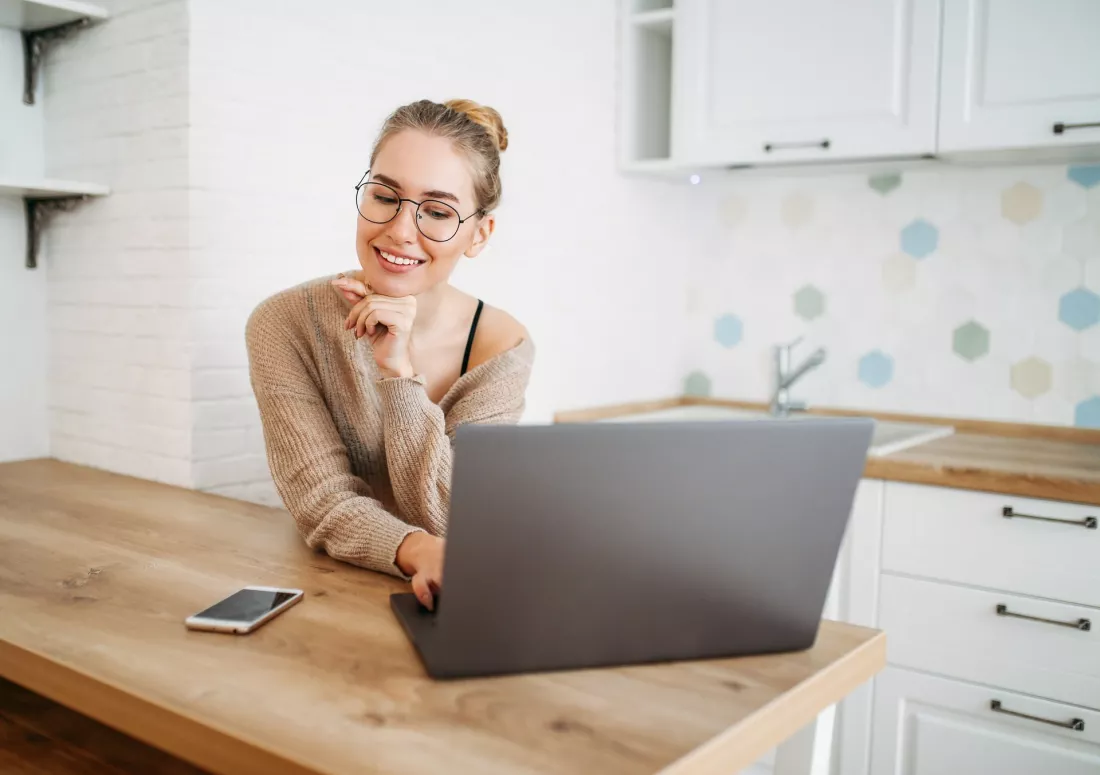 Uśmiechnięta młoda kobieta przegląda oferty na laptopie przy stole w kuchni