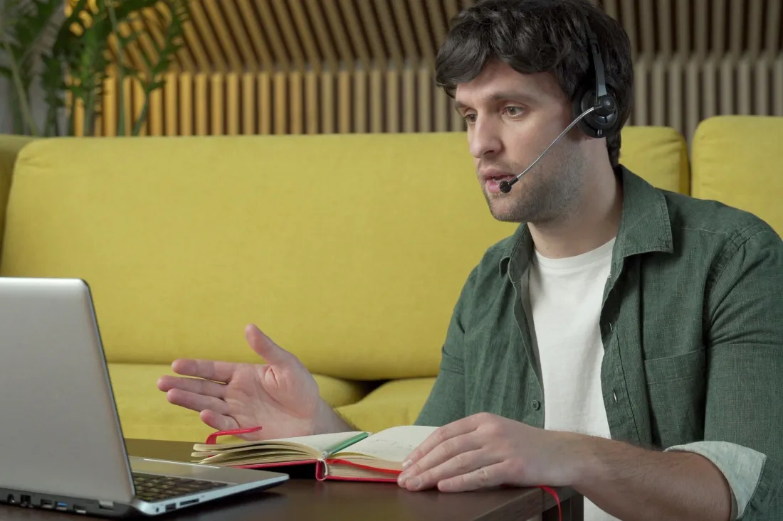 Młody mężczyzna w słuchawkach z mikrofonem rozmawia przez wideo na laptopie w domu